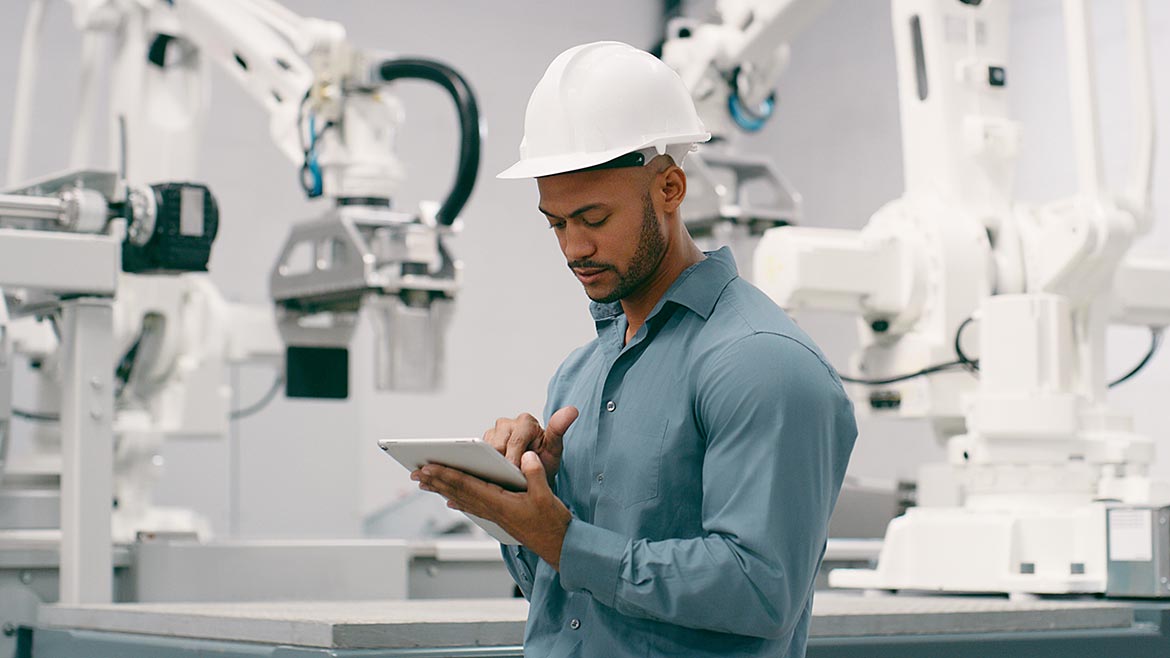 Man in factory working on a tablet with robots in the background.